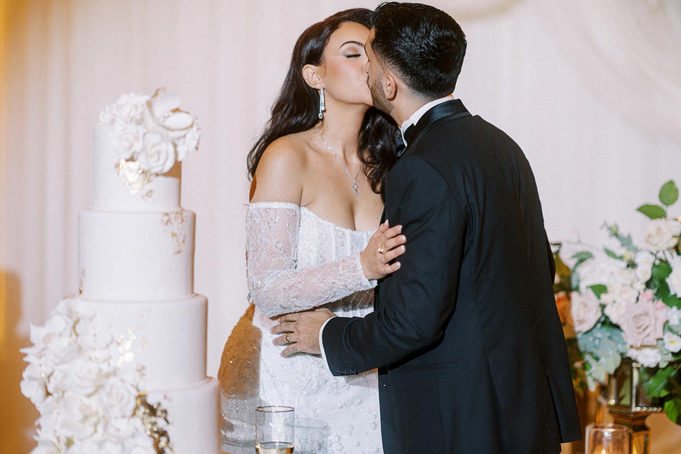 A bride and groom share a kiss by a four-tiered wedding cake adorned with floral decorations, set against a backdrop of