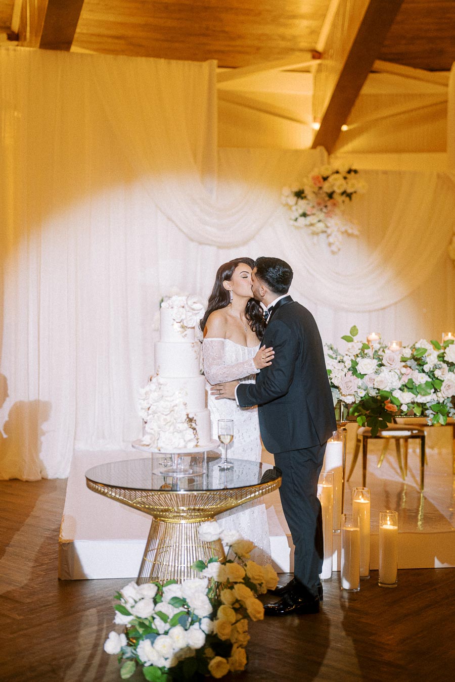 A newlywed couple shares a romantic kiss by their elegant wedding cake in a beautifully decorated reception hall, featuring