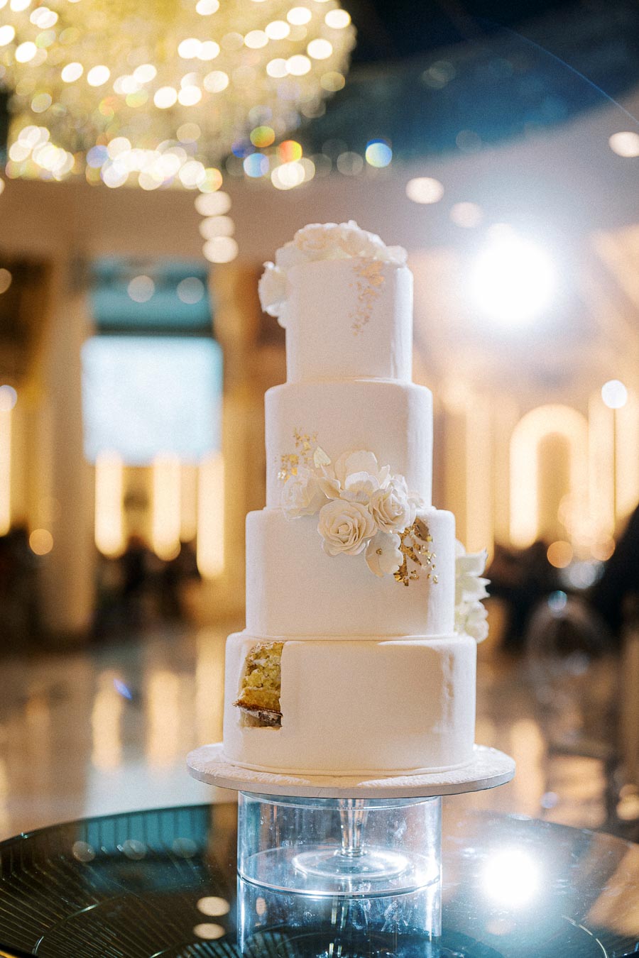 Four-tier white wedding cake adorned with elegant floral decorations and a small slice cut out, displayed on a reflective