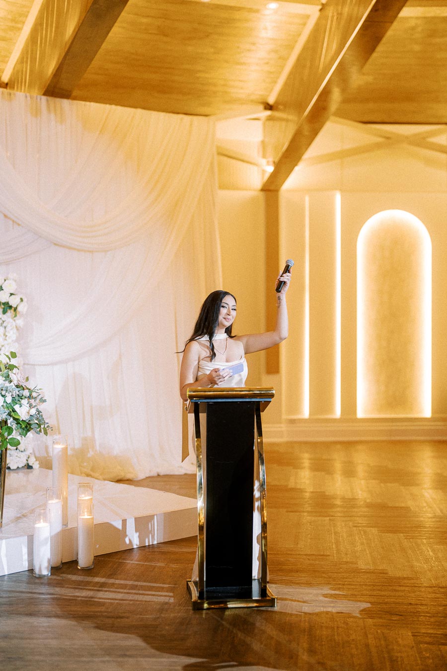 Woman giving a speech at a podium in an elegant event space with soft lighting and white floral decor.