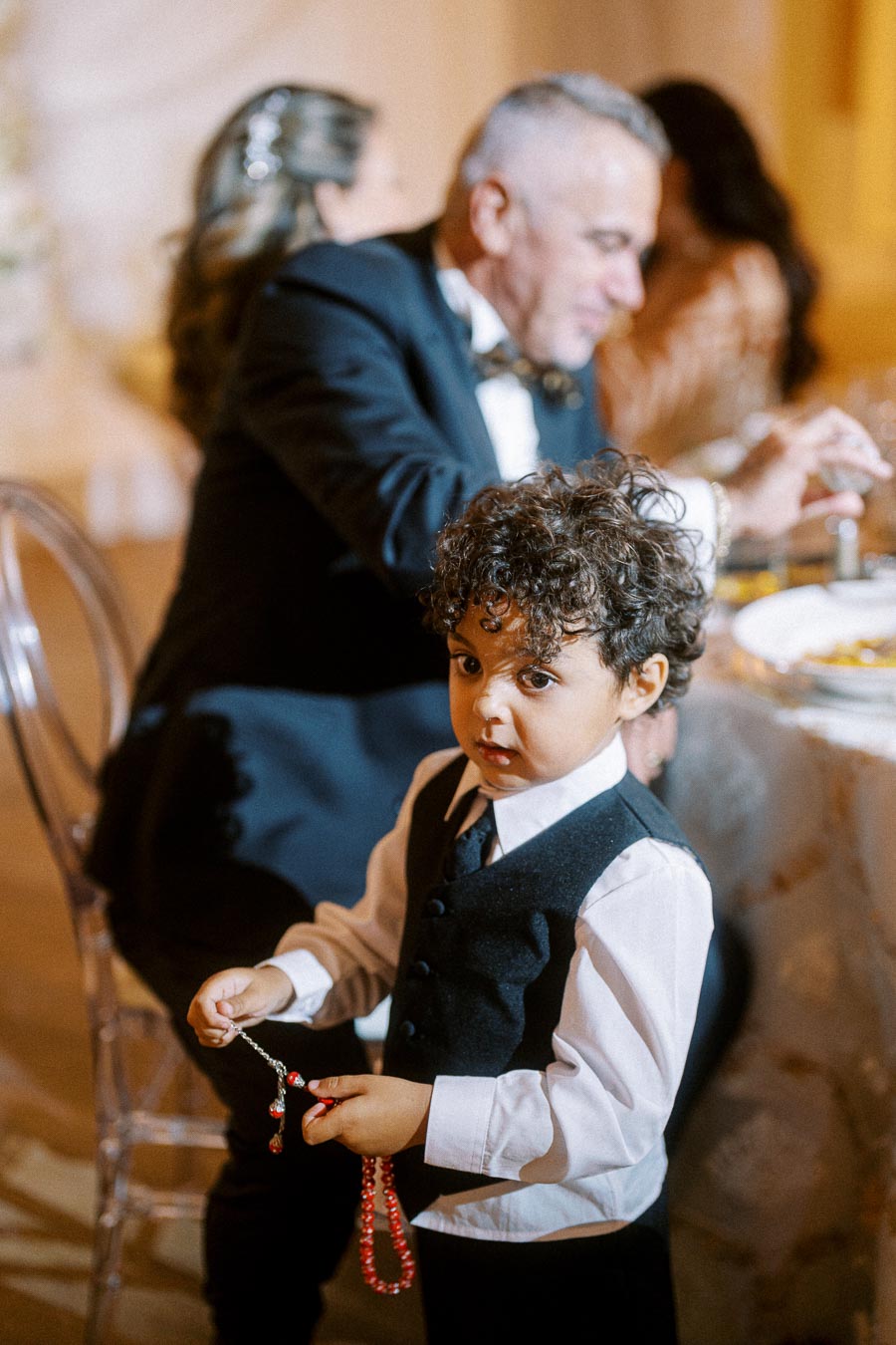 A young boy in formal attire stands holding a rosary at an event, with adults seated at a dining table in the background.