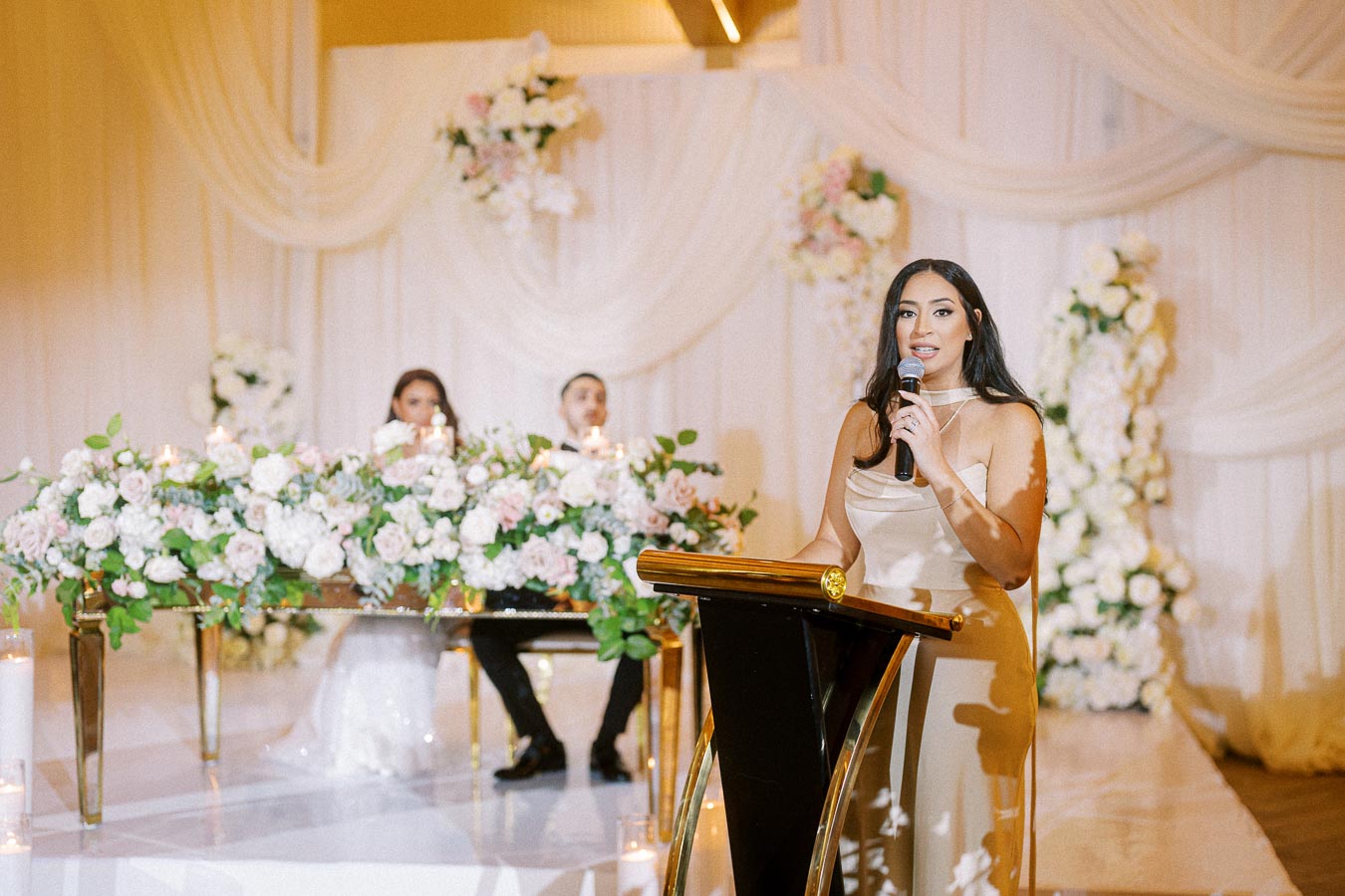 A woman in a beige dress speaks into a microphone at a beautifully decorated wedding venue. Elegant floral arrangements and