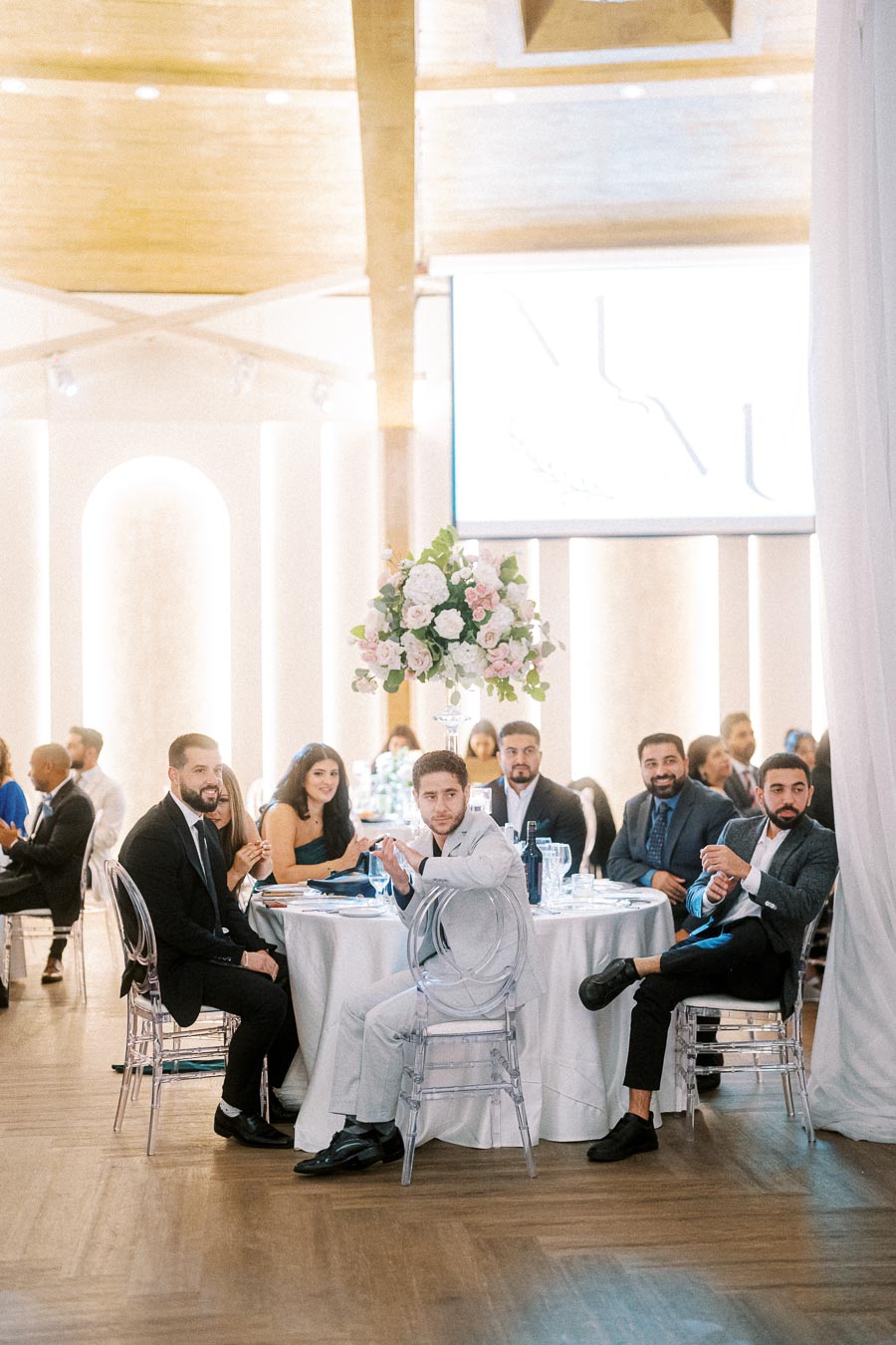 Guests sitting at a elegantly decorated table in a modern wedding reception venue, featuring white tablecloths, clear