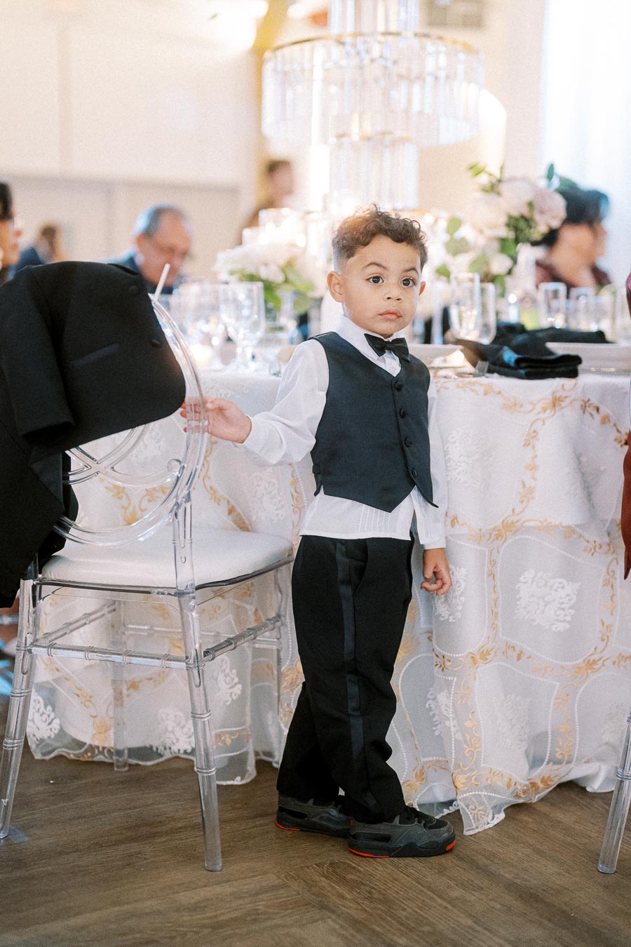 A young boy dressed in a formal black vest and white shirt stands next to a decorated table at an elegant event.