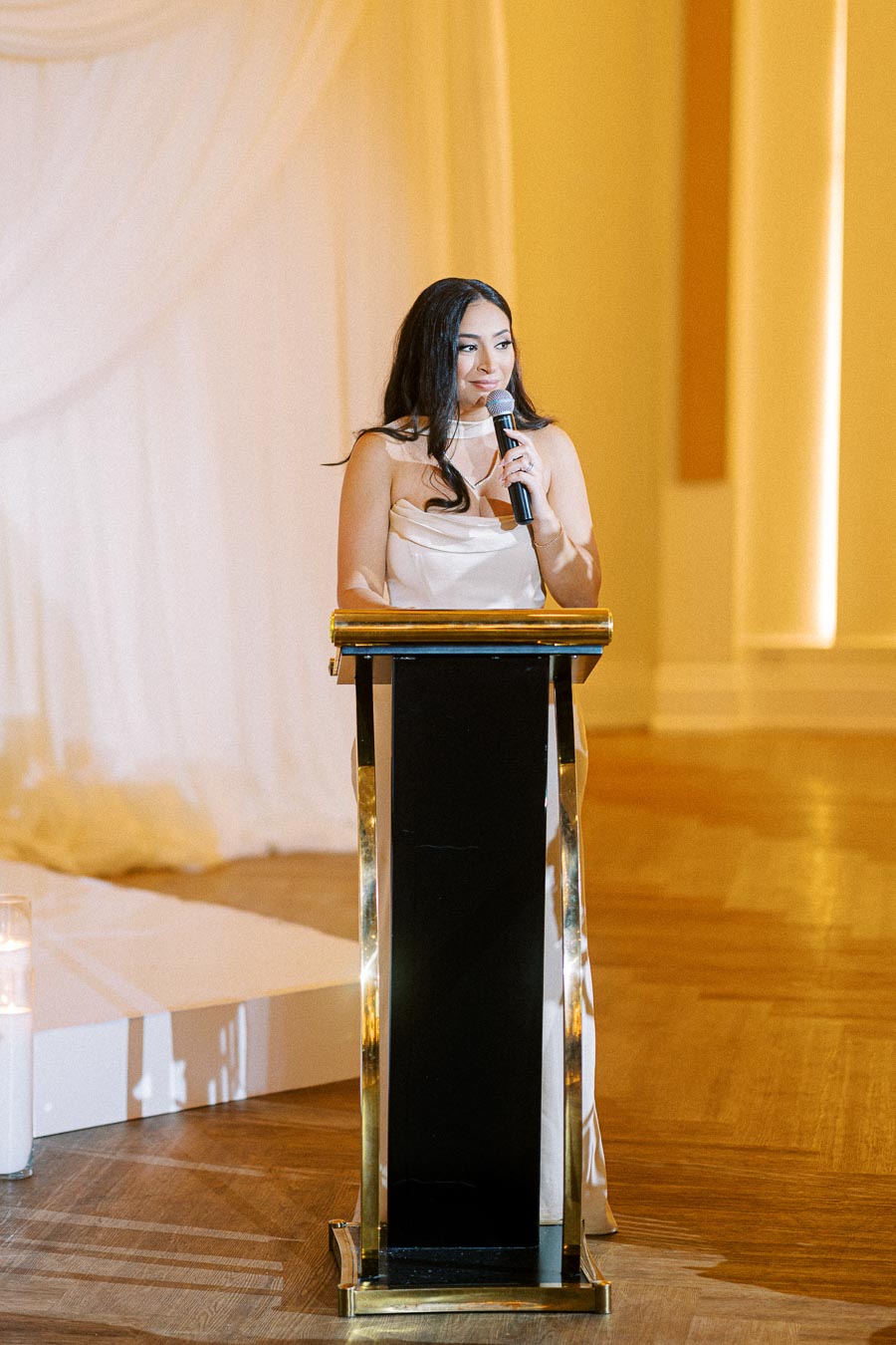 Woman in elegant dress giving a speech at a podium during an event, holding a microphone, with soft lighting