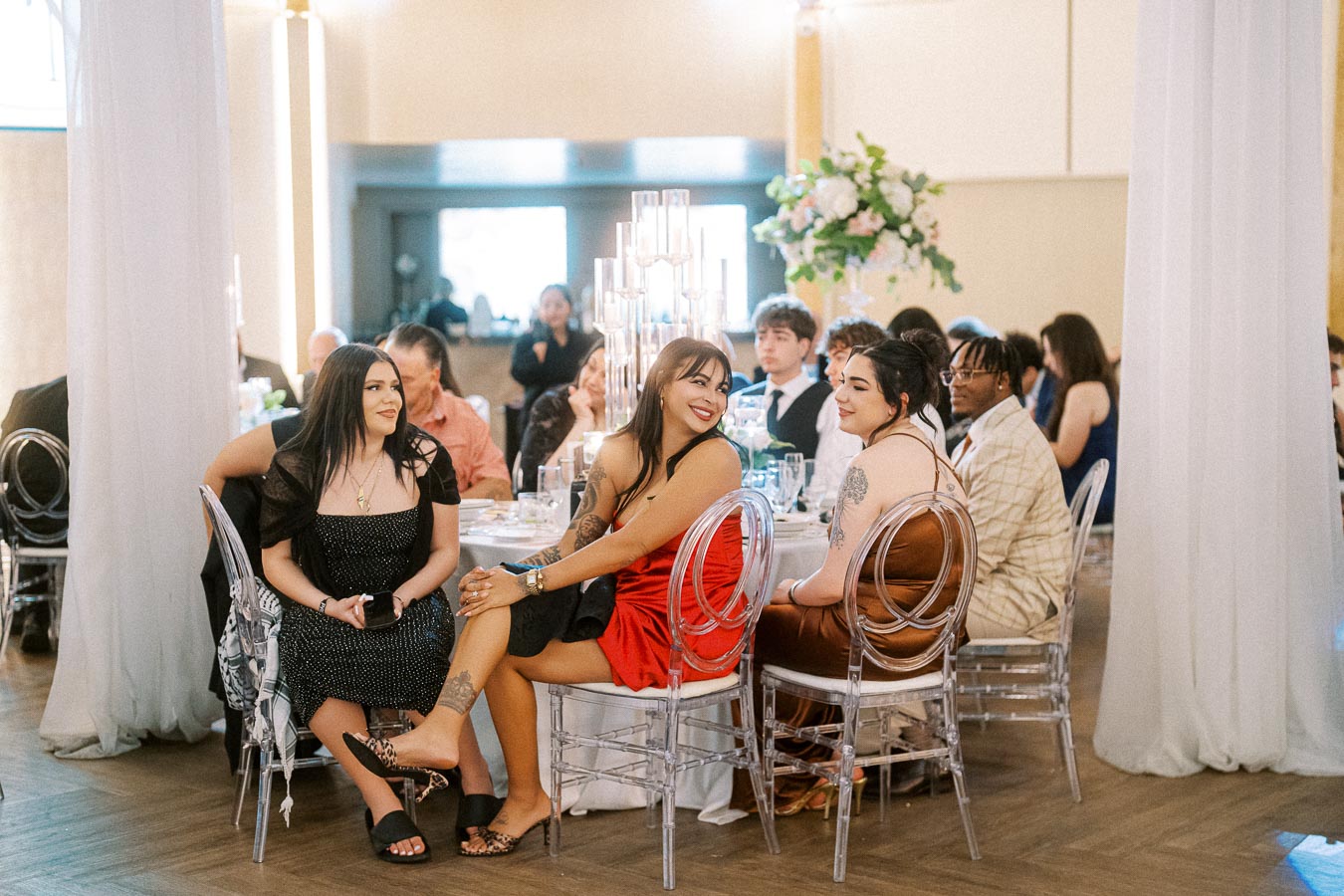 Group of elegantly dressed people enjoying a formal event, seated around a table in a beautifully decorated venue, featuring