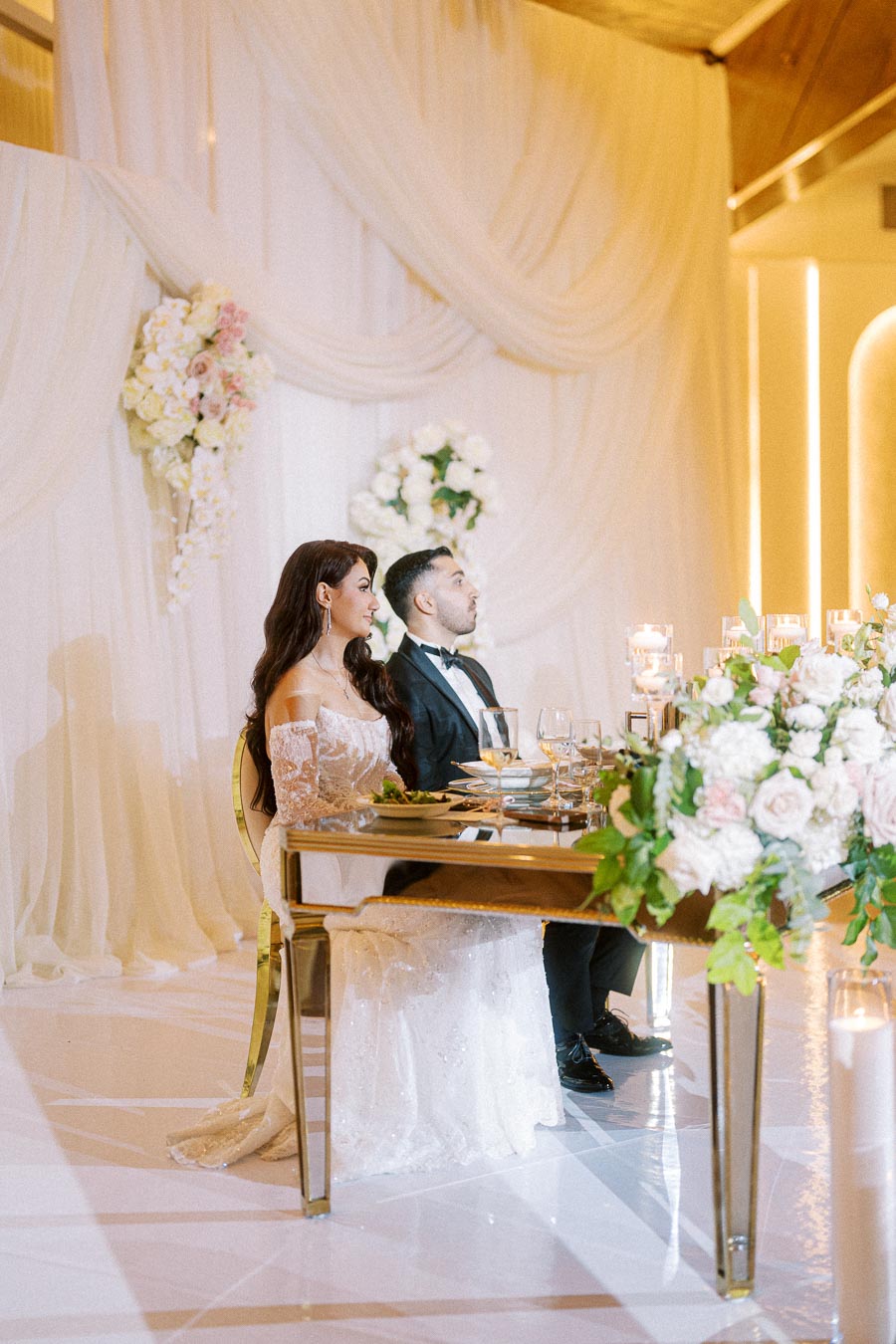 Elegant couple seated at a luxurious wedding reception, adorned with floral decorations and soft drapery, creating a