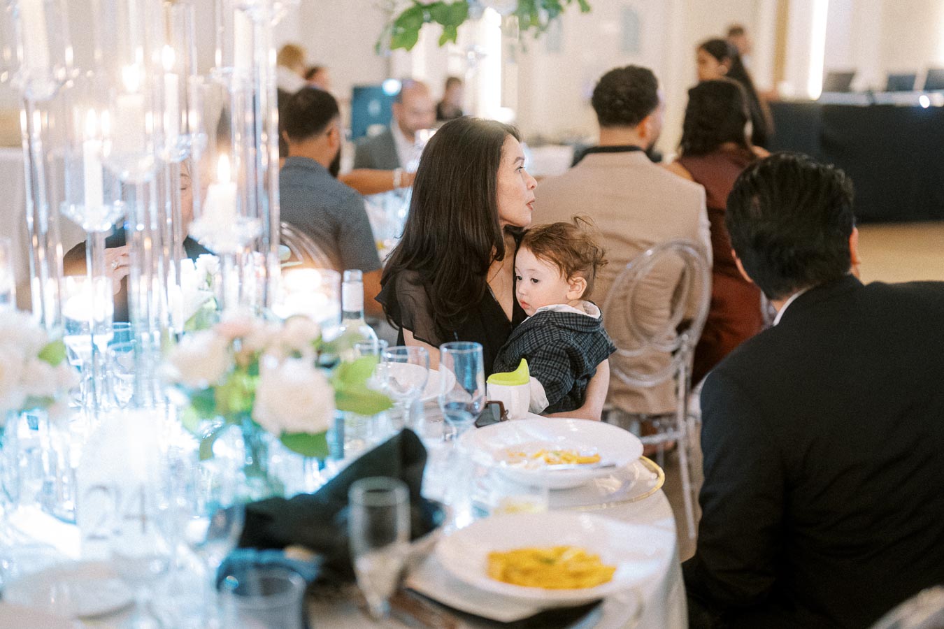 A well-dressed woman holding a child at an elegant dinner event, surrounded by candlelit tables adorned with flowers.