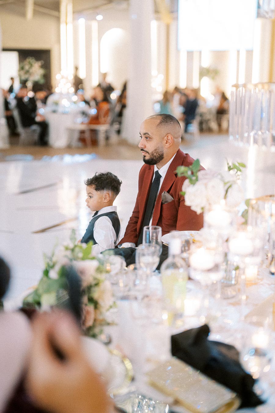 Father and son sitting at an elegant wedding reception, surrounded by beautifully arranged flowers and candles, with guests