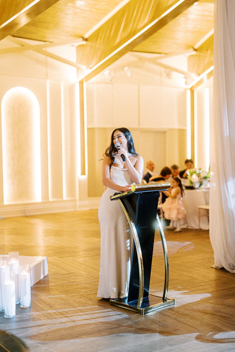 Elegant woman in a formal dress speaking into a microphone at a sophisticated indoor event with stylish decor and ambient