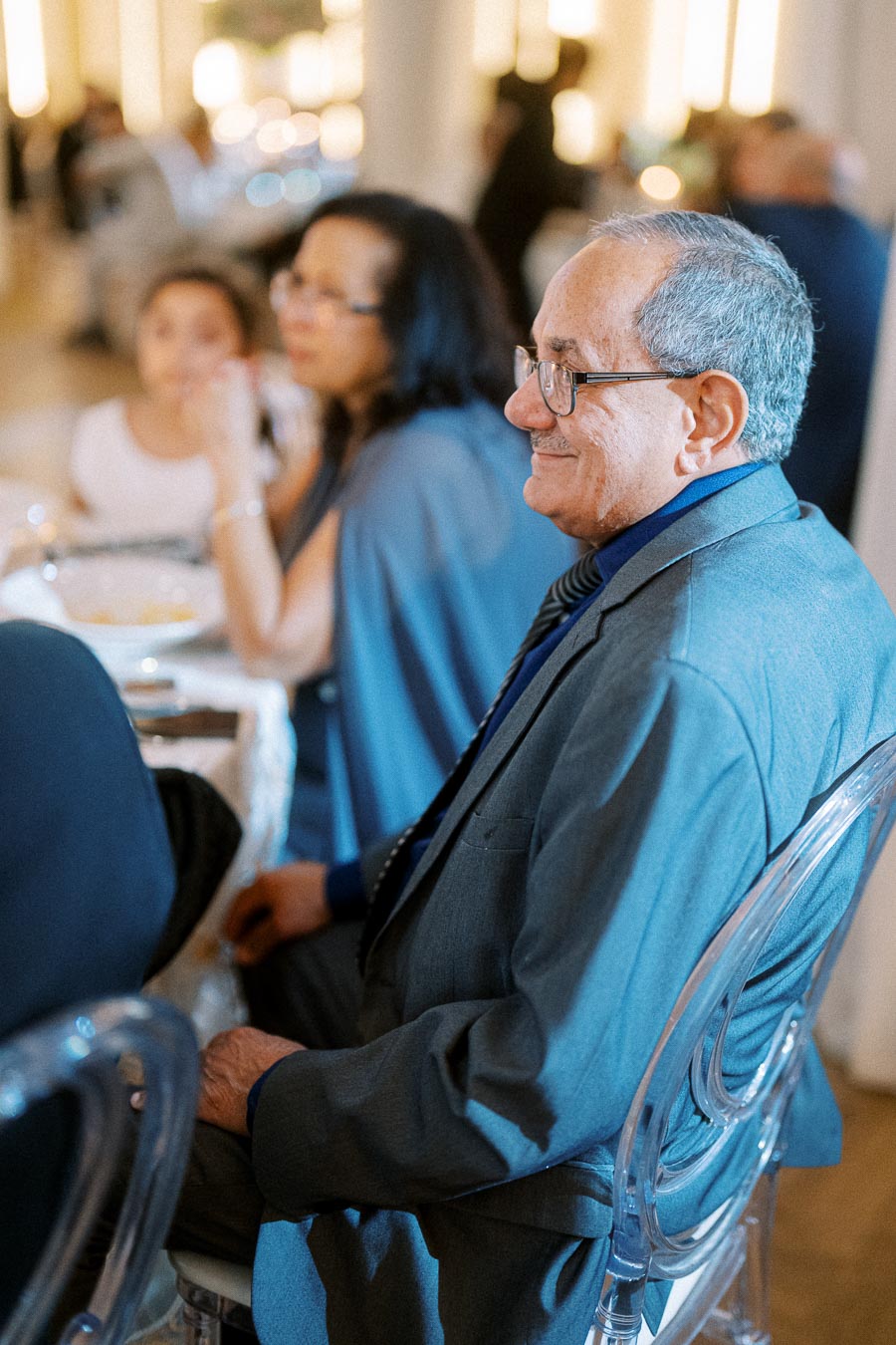 Senior man smiling at a formal event, seated at a table with blurred guests in the background, wearing a suit and glasses.