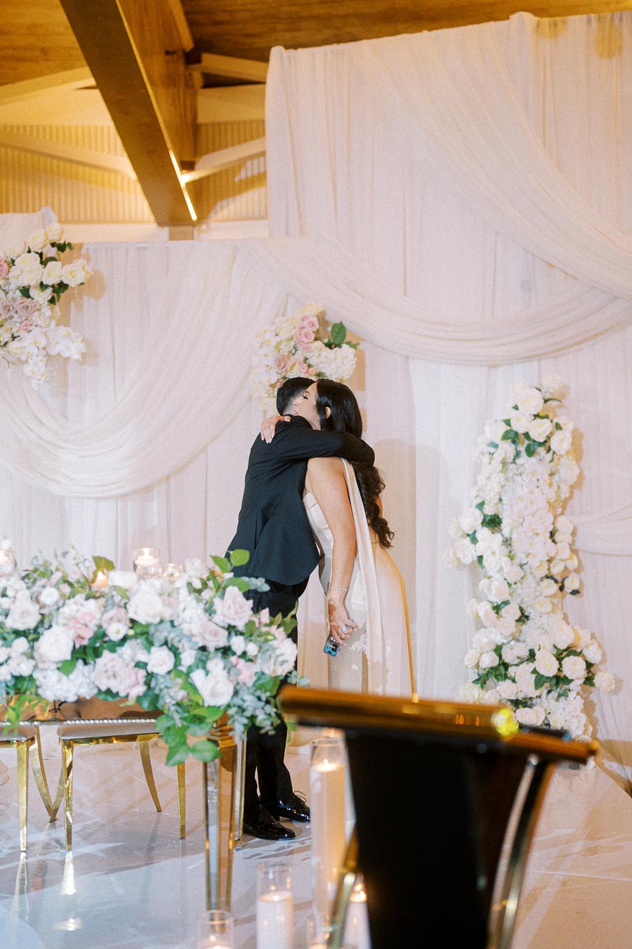 A couple embracing at a beautifully decorated wedding venue, surrounded by elegant floral arrangements and white drapery,