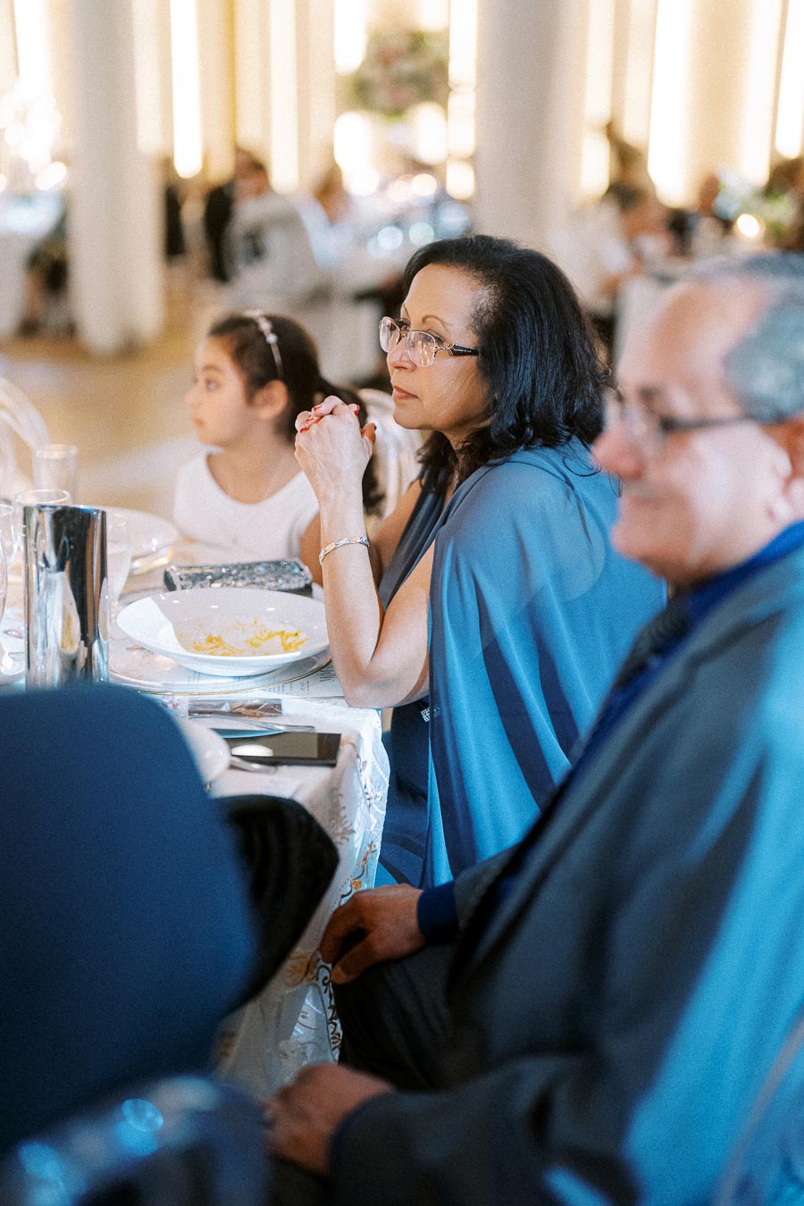 Elegant woman in glasses and blue dress attentively listening at a formal event, seated beside a young girl in a bustling