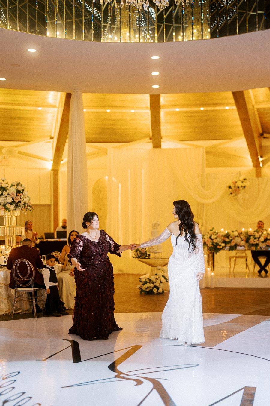 A bride and her mother share a dance on the ballroom floor at a wedding reception. The bride is wearing a white gown while