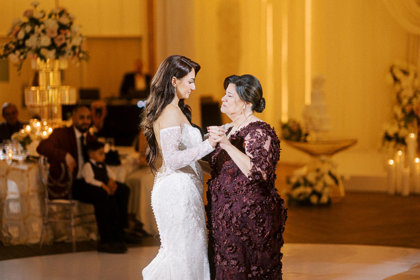 A bride in a white wedding dress shares a joyful dance with an older woman in a maroon dress at a wedding reception,