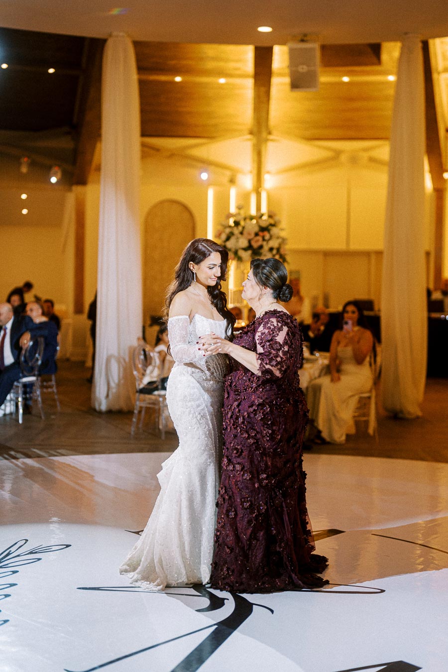 A bride in an elegant white gown dances with an older woman in a burgundy dress during a wedding reception, creating a