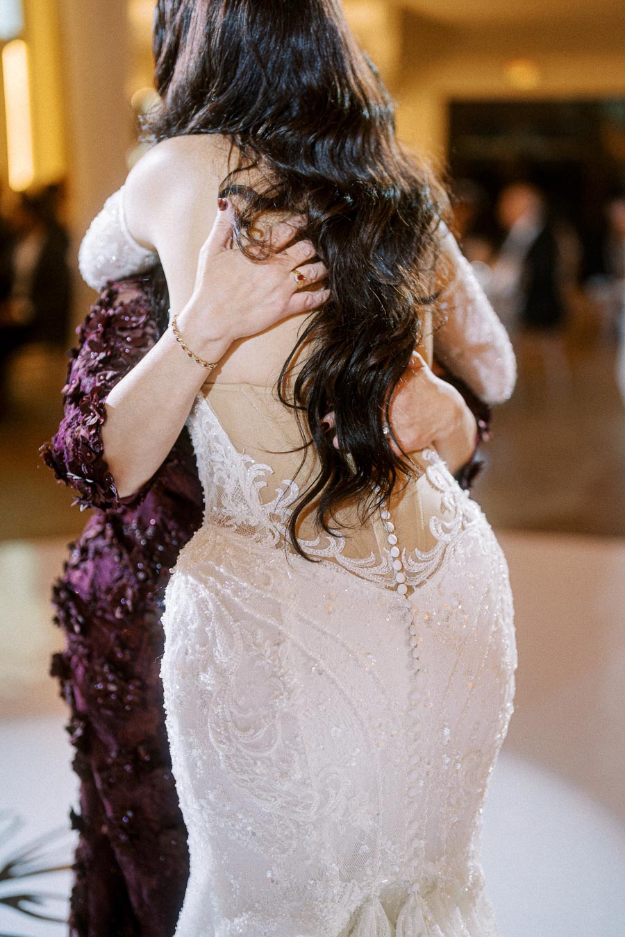 A close-up of two individuals embracing at a wedding, with focus on the intricate details of the bride's lace gown and long,