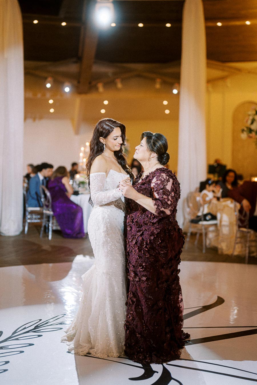 A bride in a white lace wedding dress shares a heartfelt dance with a woman in an elegant burgundy gown at a warmly lit