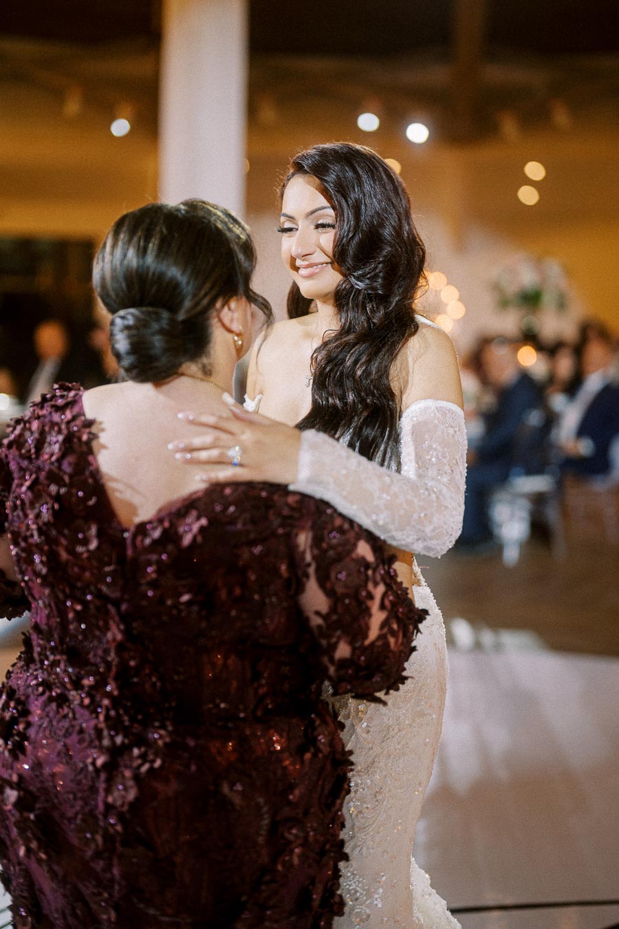 Bride in elegant lace wedding dress smiling and embracing a woman in a burgundy gown during a wedding celebration.