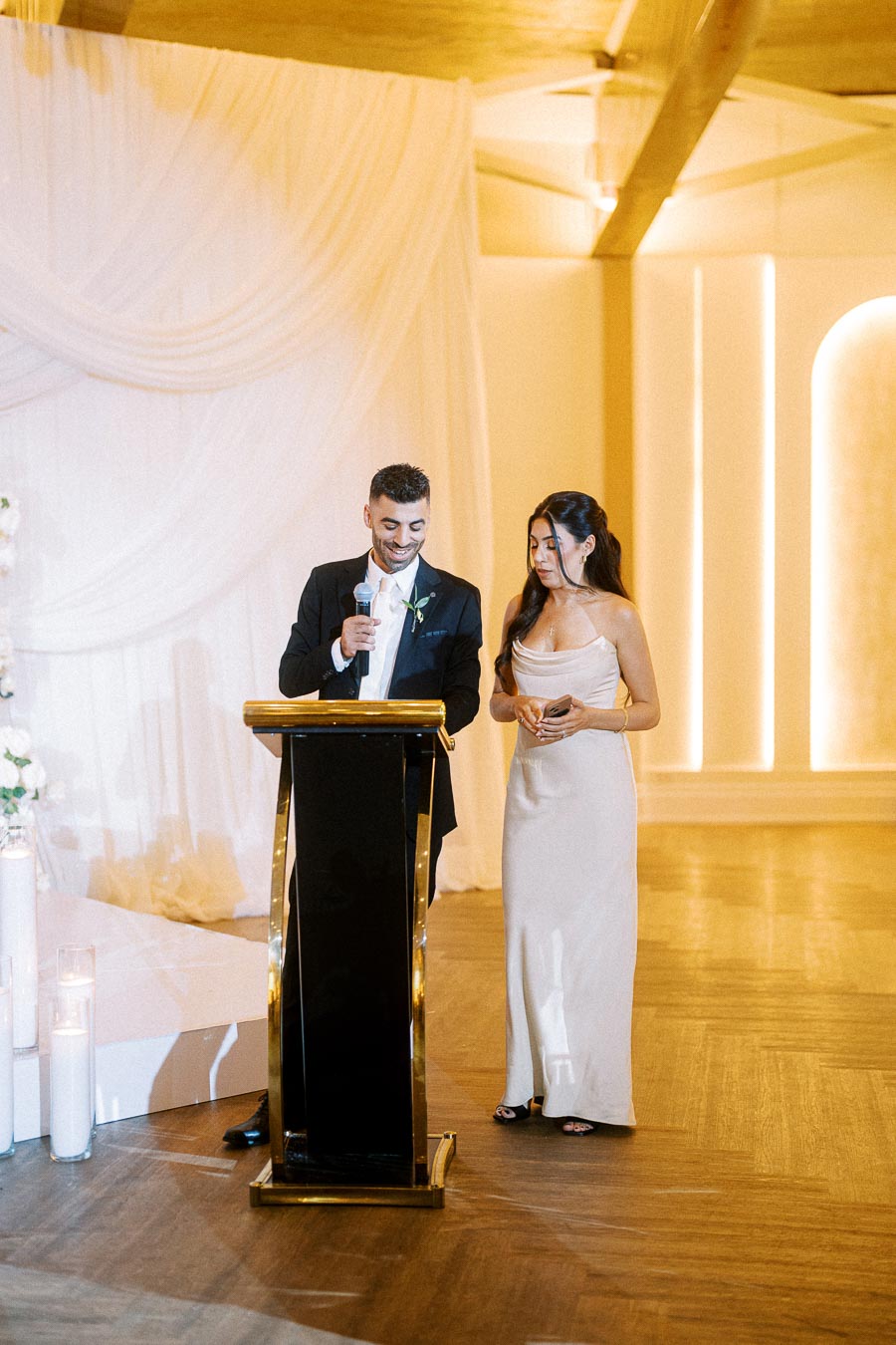 Two elegantly dressed individuals delivering a speech at a formal event, standing behind a podium in a warmly lit venue.