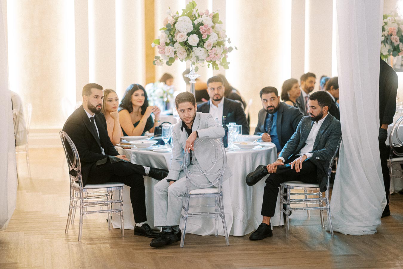 Guests seated at a wedding reception, elegantly dressed men and women around a table with decorative floral arrangements, in