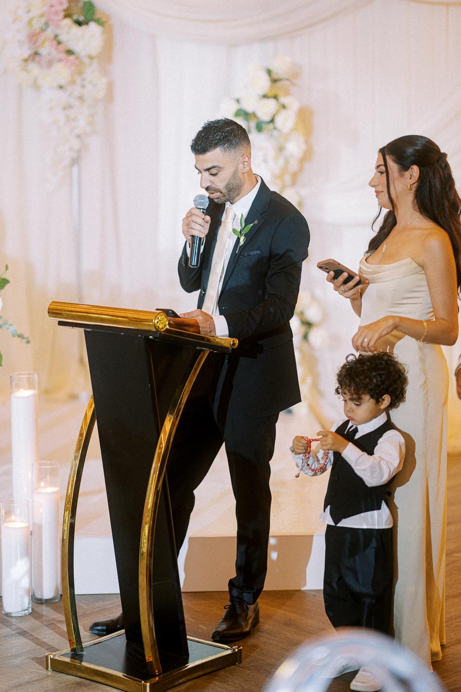 A man in a suit giving a speech with a microphone at a formal event, accompanied by a woman in an elegant dress holding a