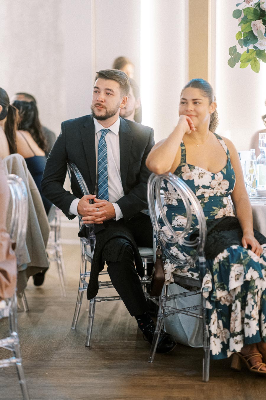Guests seated at an elegant event, featuring a man in a suit and tie and a woman in a floral dress, sitting on transparent
