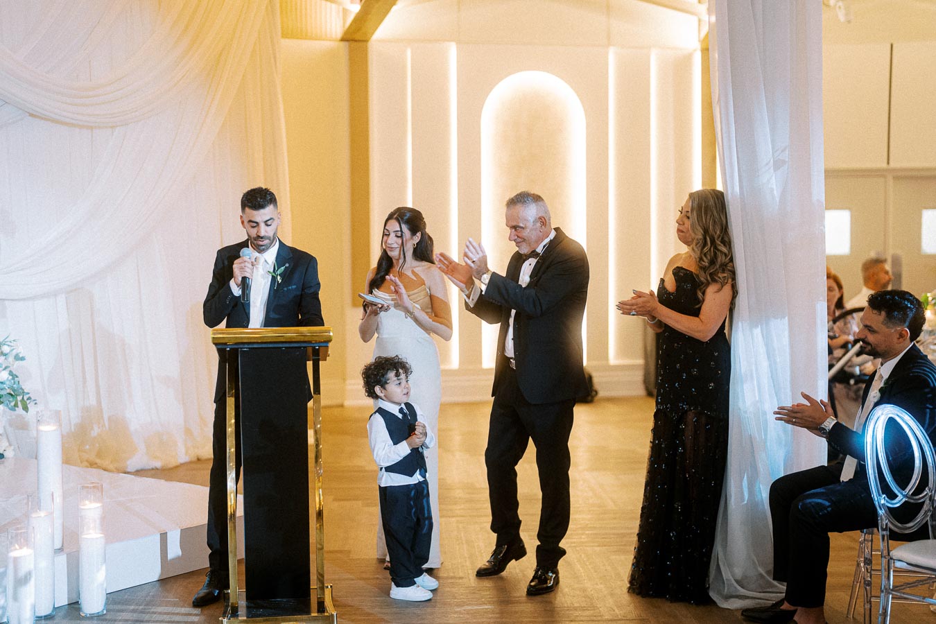 Wedding ceremony speech at an elegant venue with a well-dressed man speaking at a podium. A woman in a white dress and other