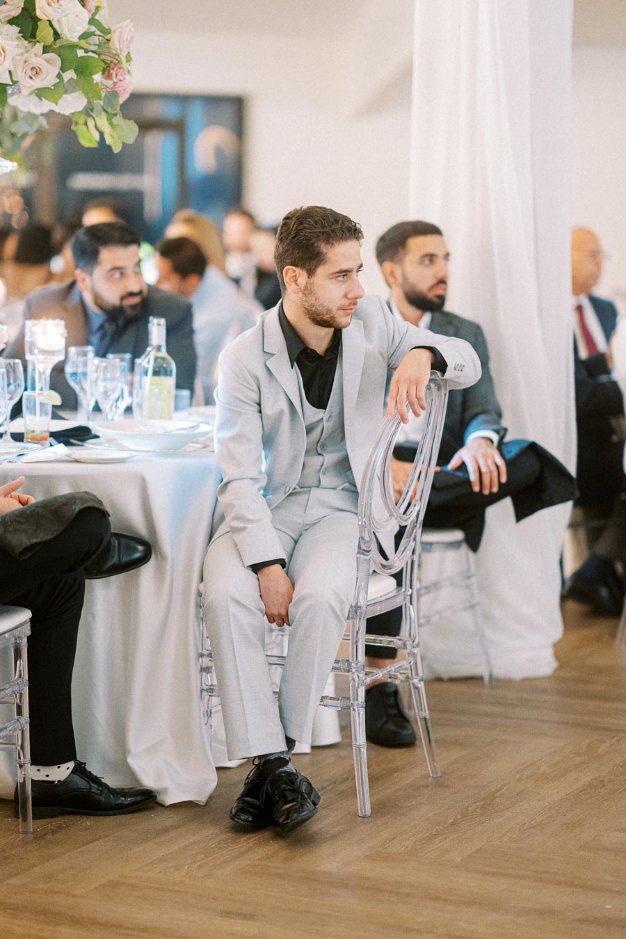 Man in a light gray suit sitting on a clear chair at an elegant event, surrounded by people in formal attire, with a