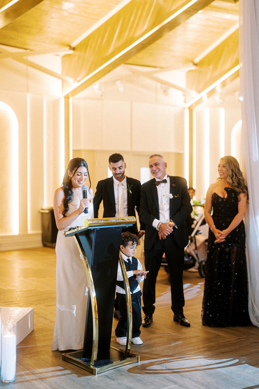 A group of people dressed formally, standing at a podium with a microphone in a well-lit event space, possibly giving a