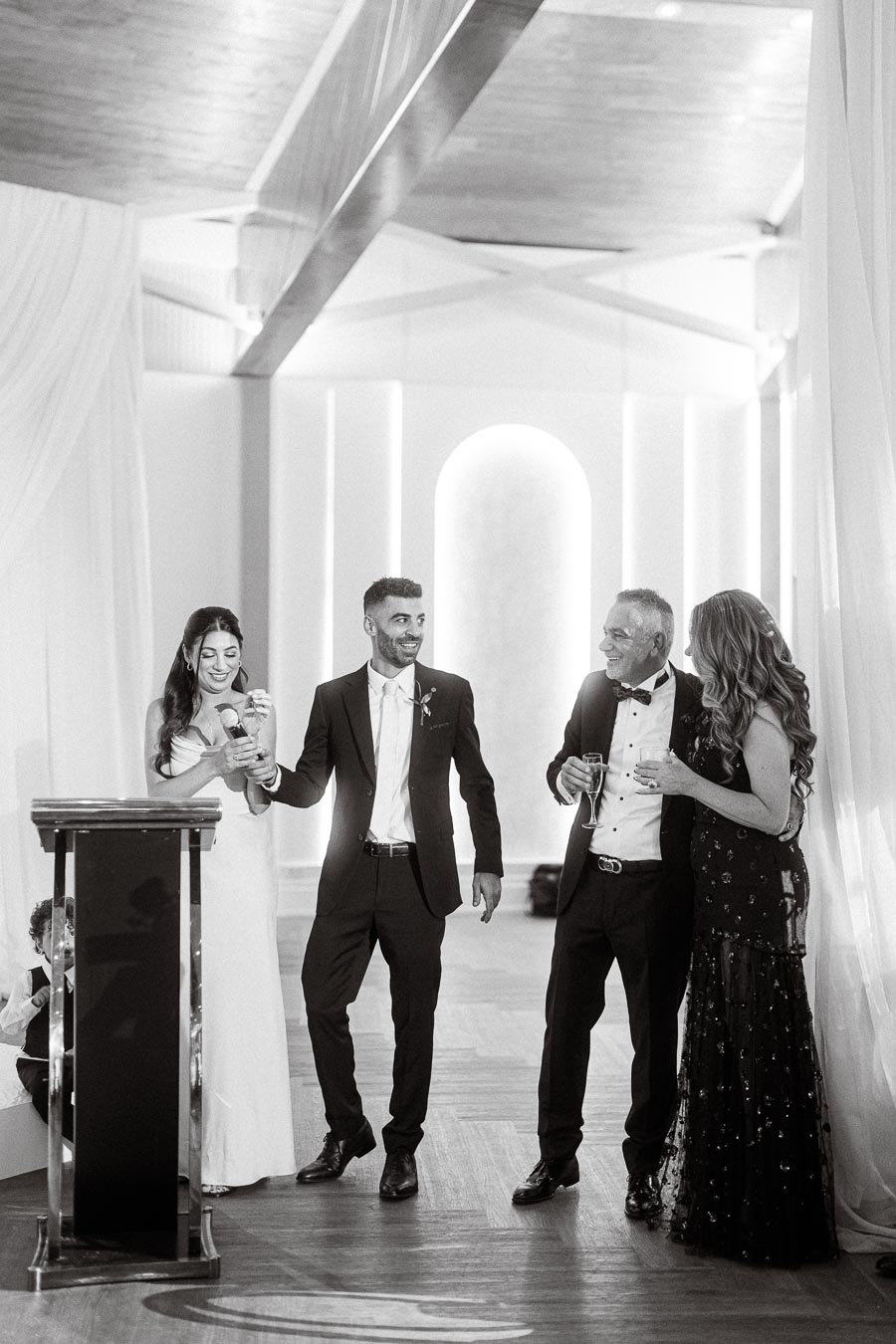 A black and white photo of four elegantly dressed people standing and smiling at a wedding reception. The group appears to