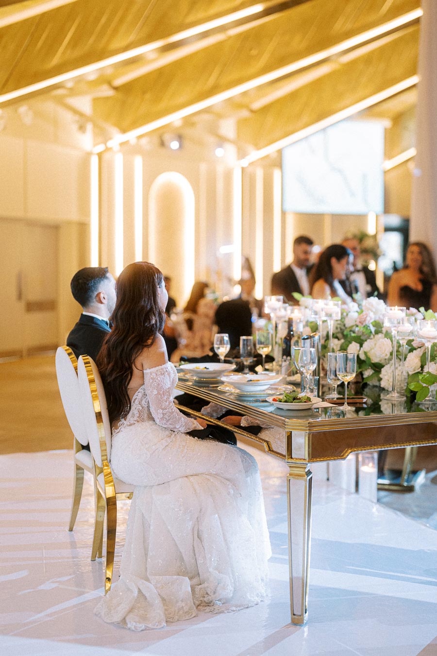 Elegant wedding reception scene with a bride in a lace gown seated beside the groom at a beautifully decorated table,