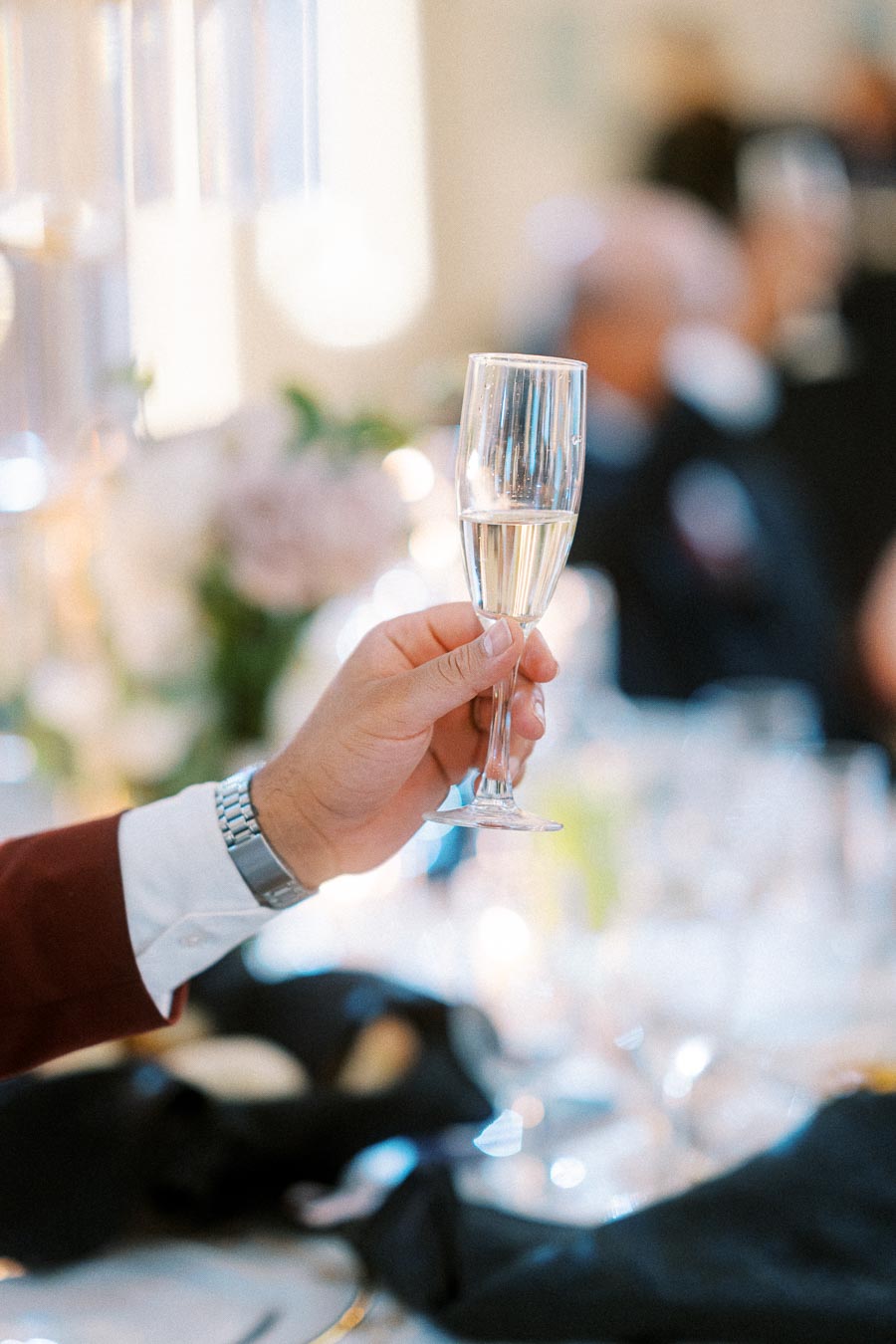 A close-up of a person holding a champagne flute in a celebratory setting, with a blurred background of elegant table