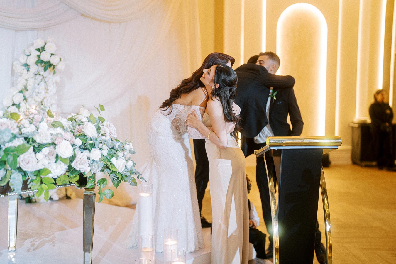 A wedding ceremony scene with two couples embracing on a stage, surrounded by elegant floral arrangements and ambient