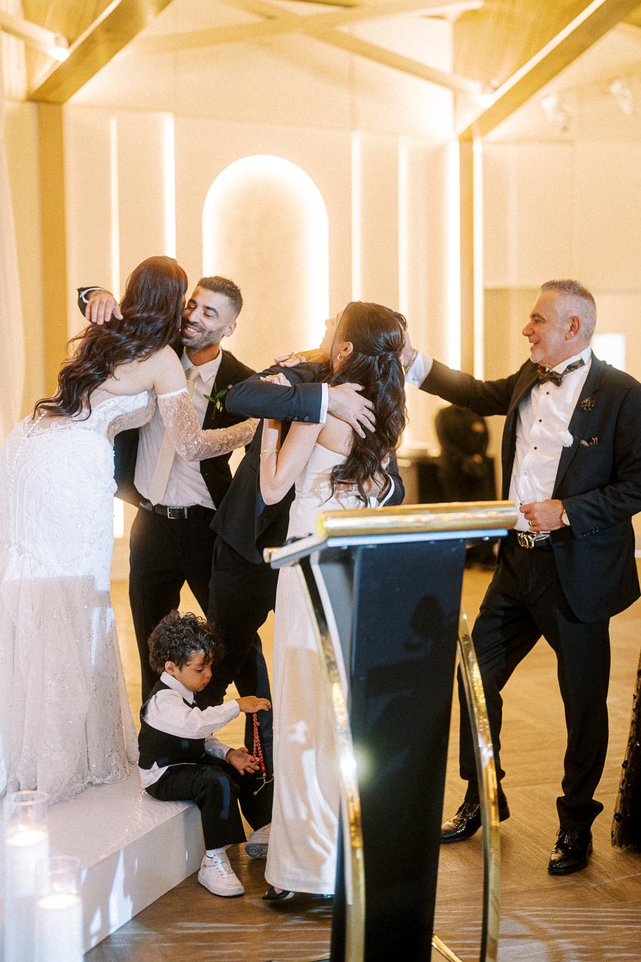 A joyful wedding celebration with a warmly lit room featuring guests in formal attire embracing and smiling, a young child