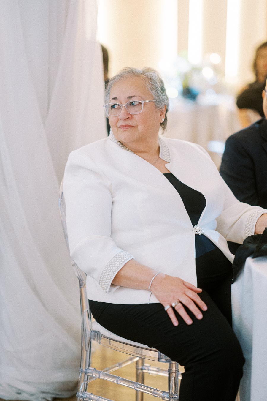 Elderly woman sitting elegantly in a white jacket at a formal event, looking to the side with a soft smile, surrounded by a