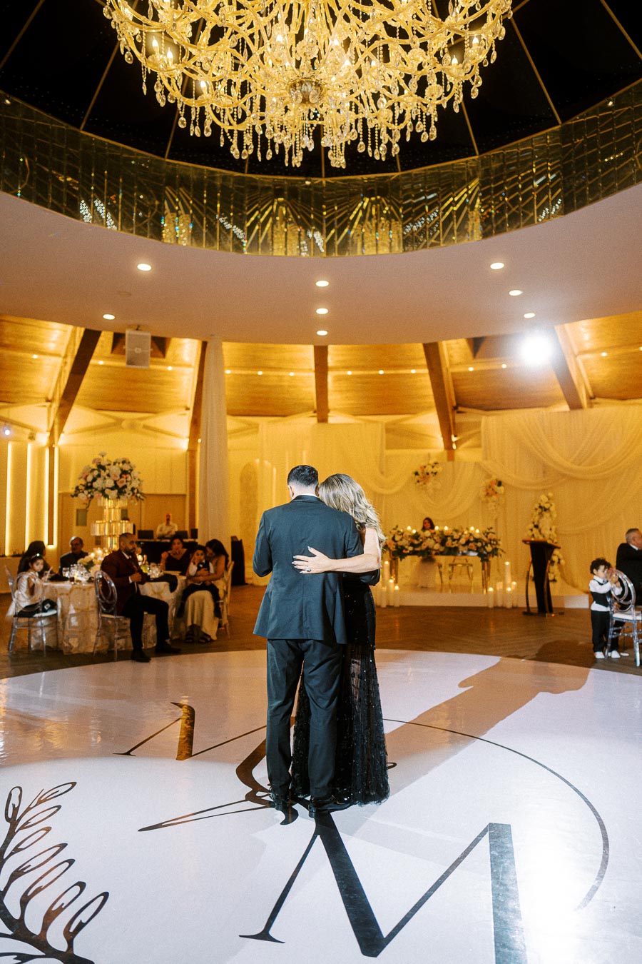 A couple shares a romantic dance under a grand chandelier in an elegant wedding venue with guests seated around them.