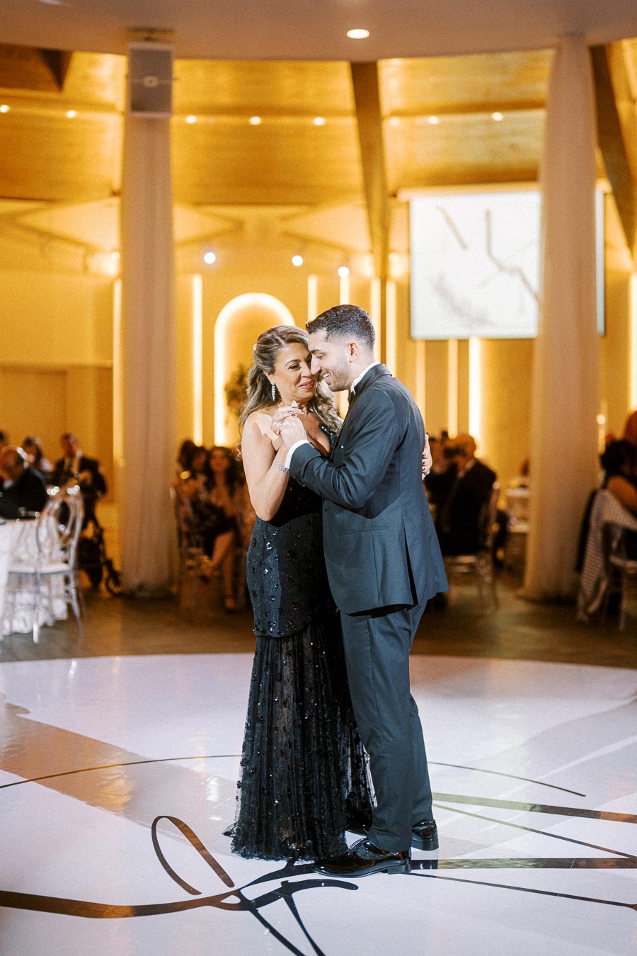 Elegant couple sharing a dance at a formal event, dressed in a black evening gown and a suit, surrounded by warmly lit