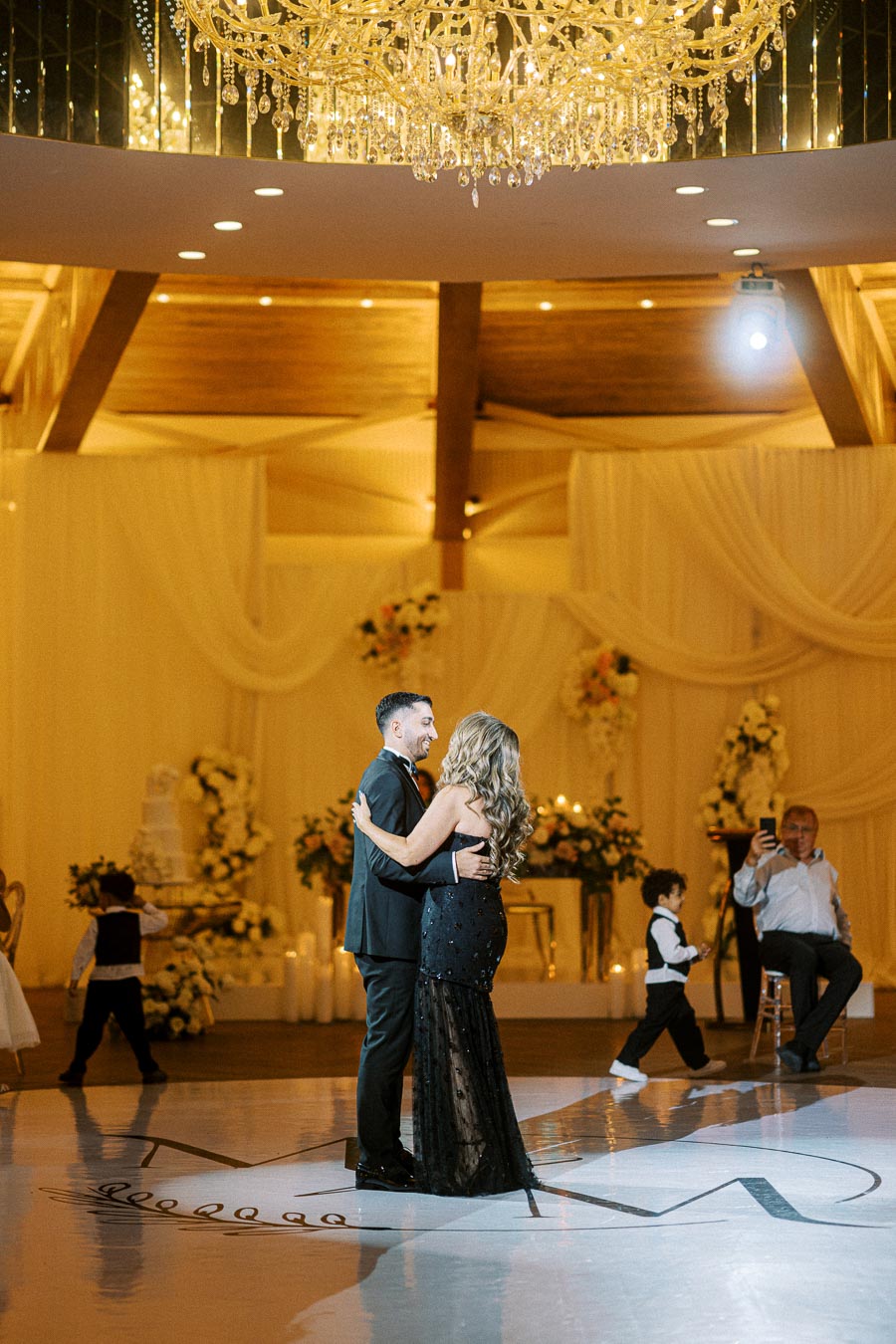 Elegant couple dancing under a luxurious chandelier at a wedding reception, surrounded by festive decorations and guests,