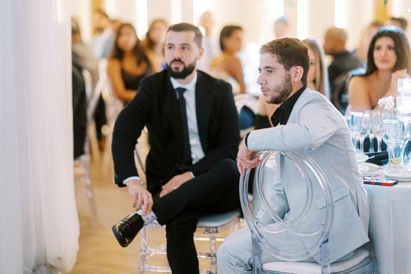 Two men in formal suits attentively watching an event at a banquet hall, with elegantly dressed guests in the background,