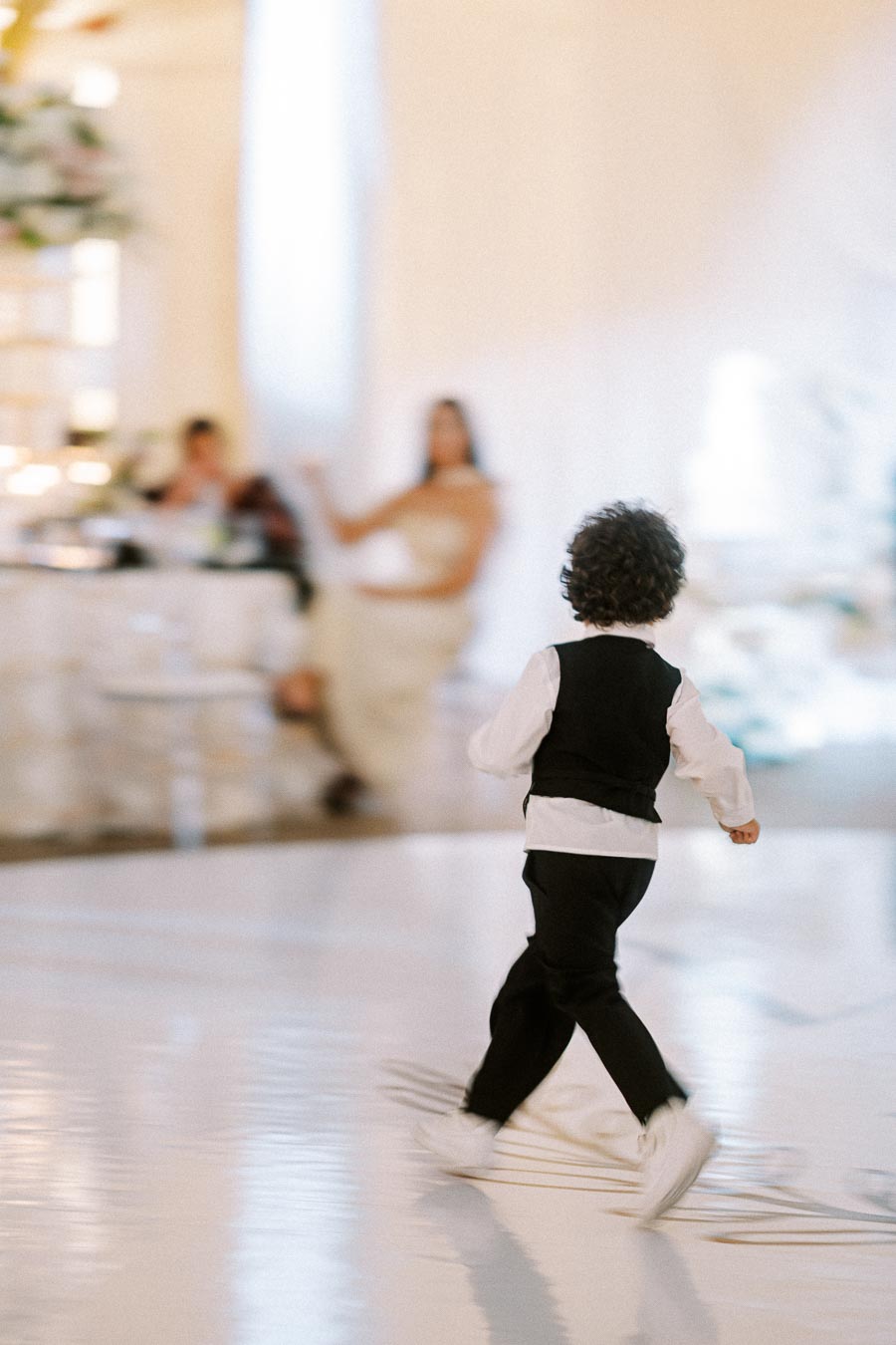 A young boy in a formal vest and pants playfully walks across a polished floor at a wedding reception, with guests seated in
