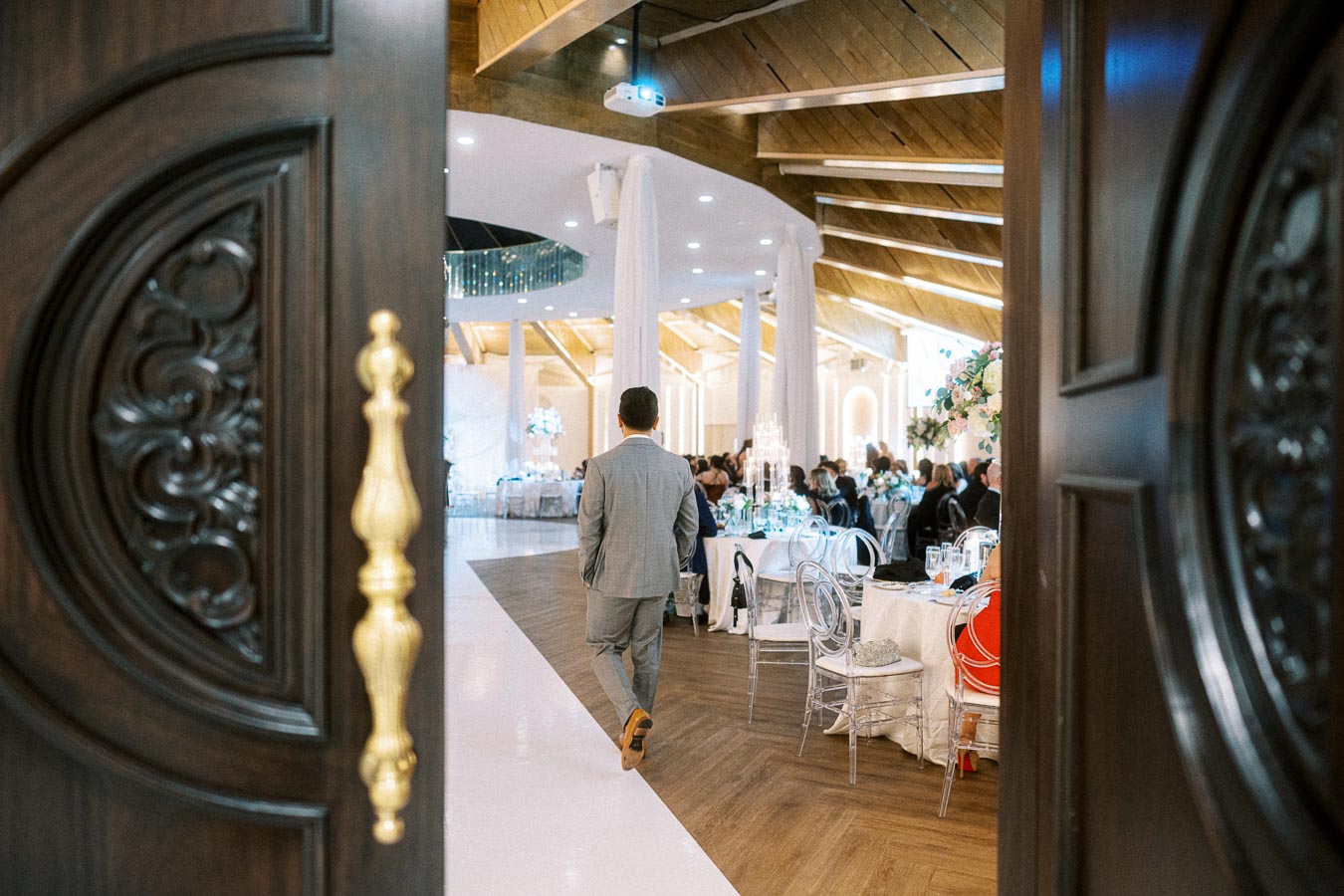 A man in a gray suit walking through ornate wooden doors into a luxurious banquet hall, featuring elegantly set tables with