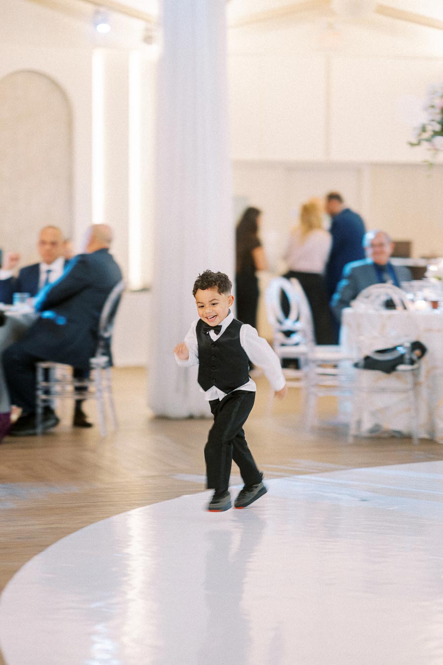 A joyful young boy in a formal outfit running across a ballroom floor, with guests seated at tables in the background of an