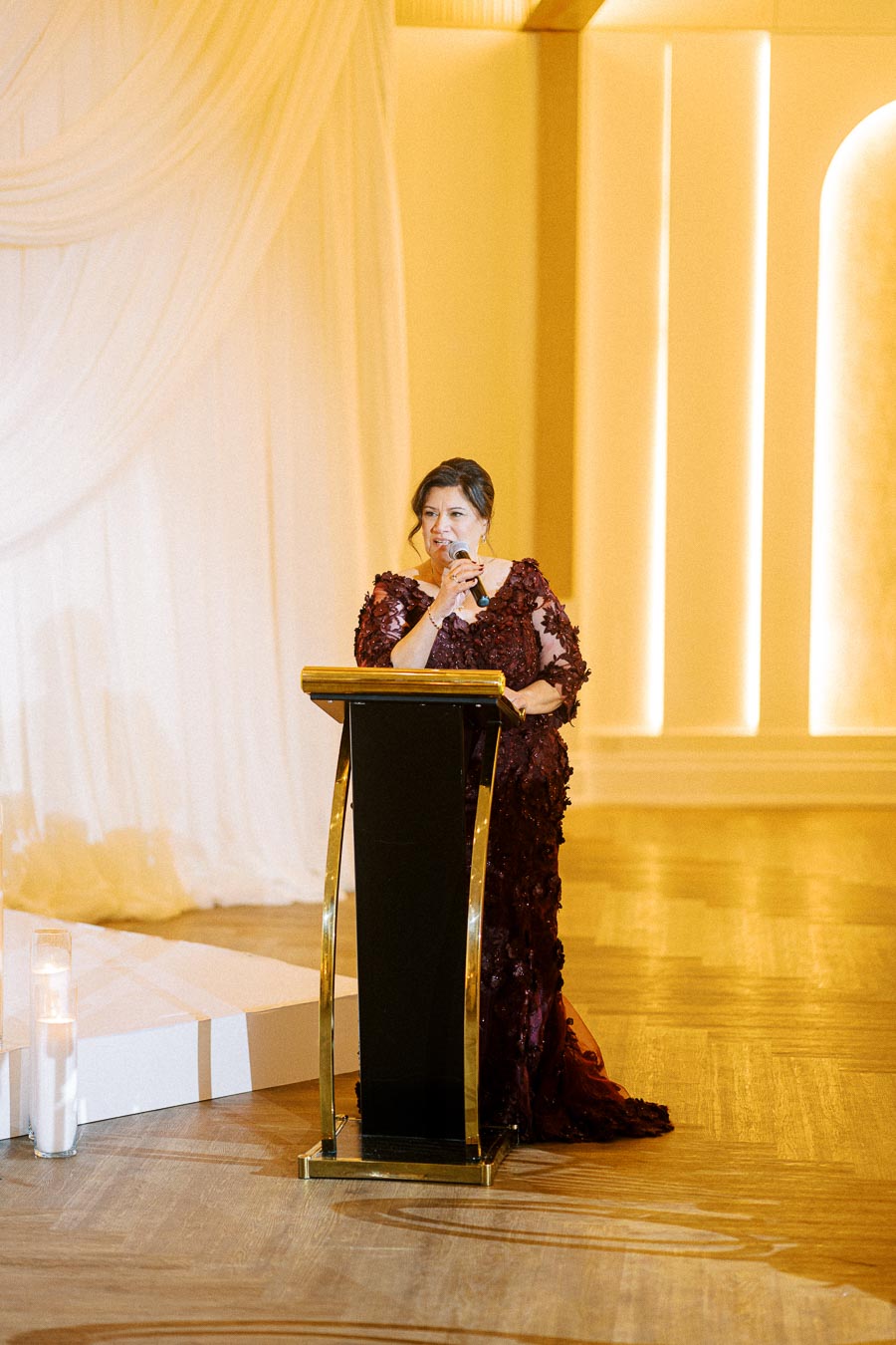 A woman in an elegant maroon dress speaking at a podium in a beautifully lit event hall.