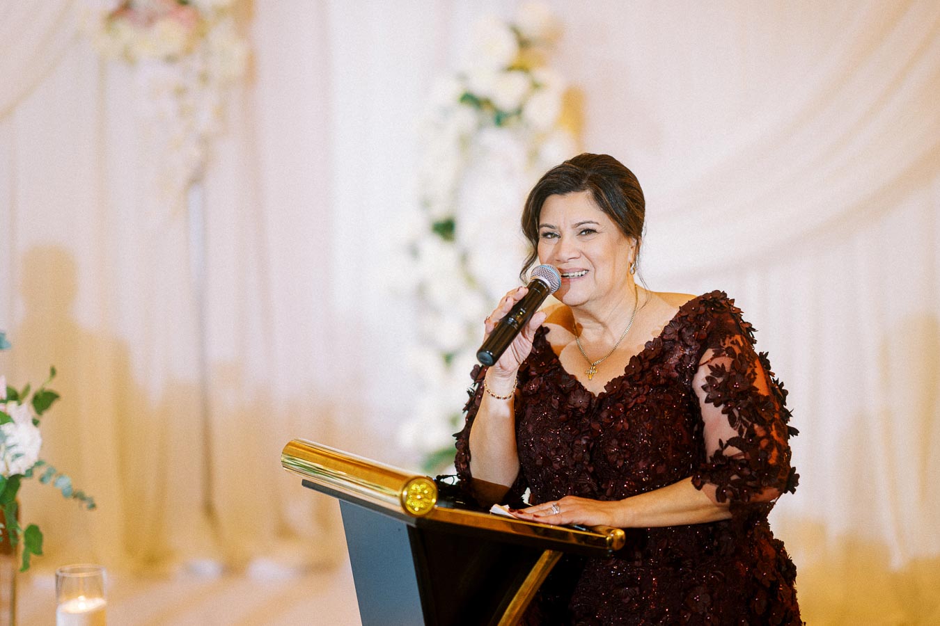 A woman in an elegant burgundy dress delivers a speech with a microphone at a formal event, standing at a podium adorned