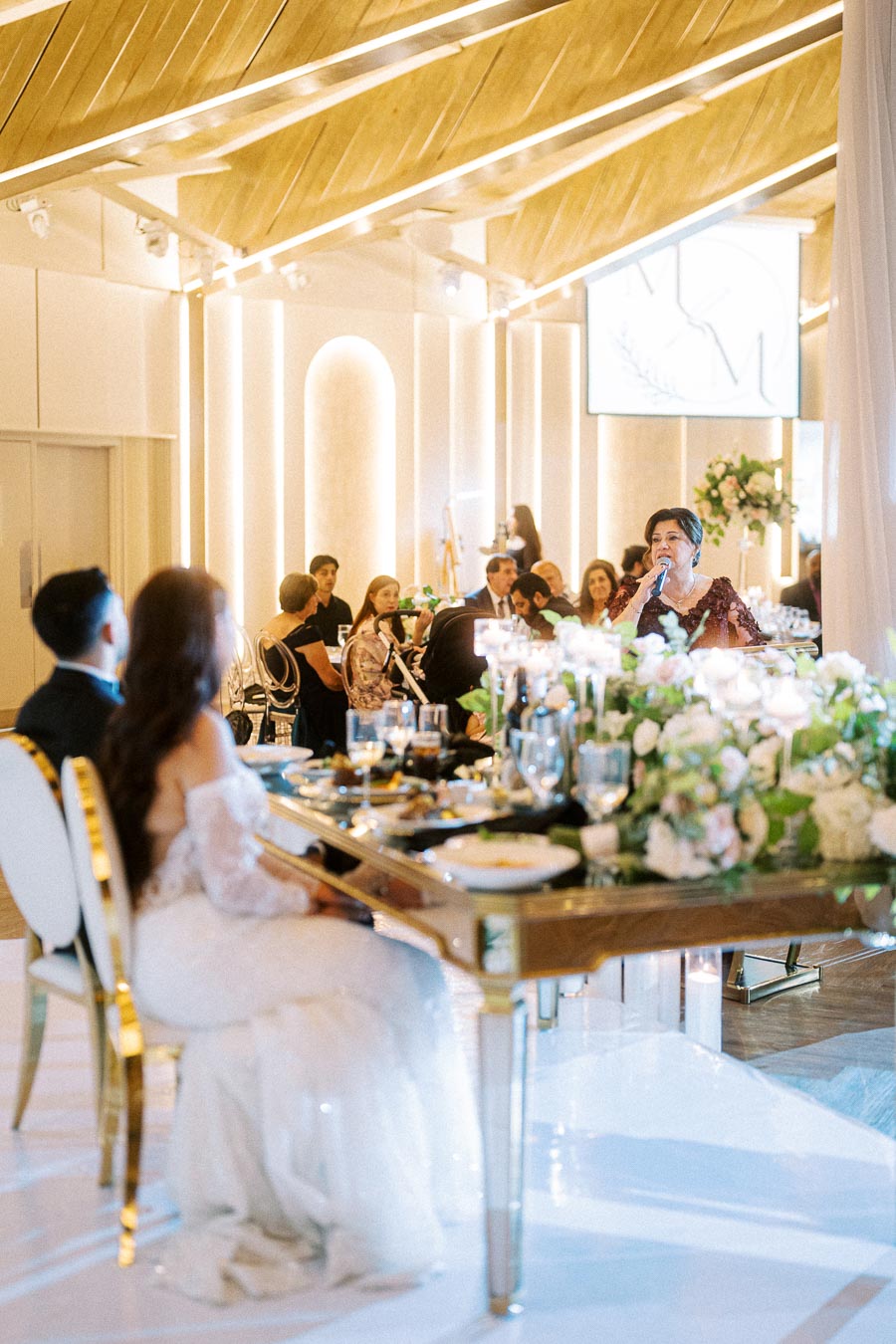 Elegant wedding reception in a modern venue with wooden ceiling and ambient lighting, featuring a woman giving a speech at a