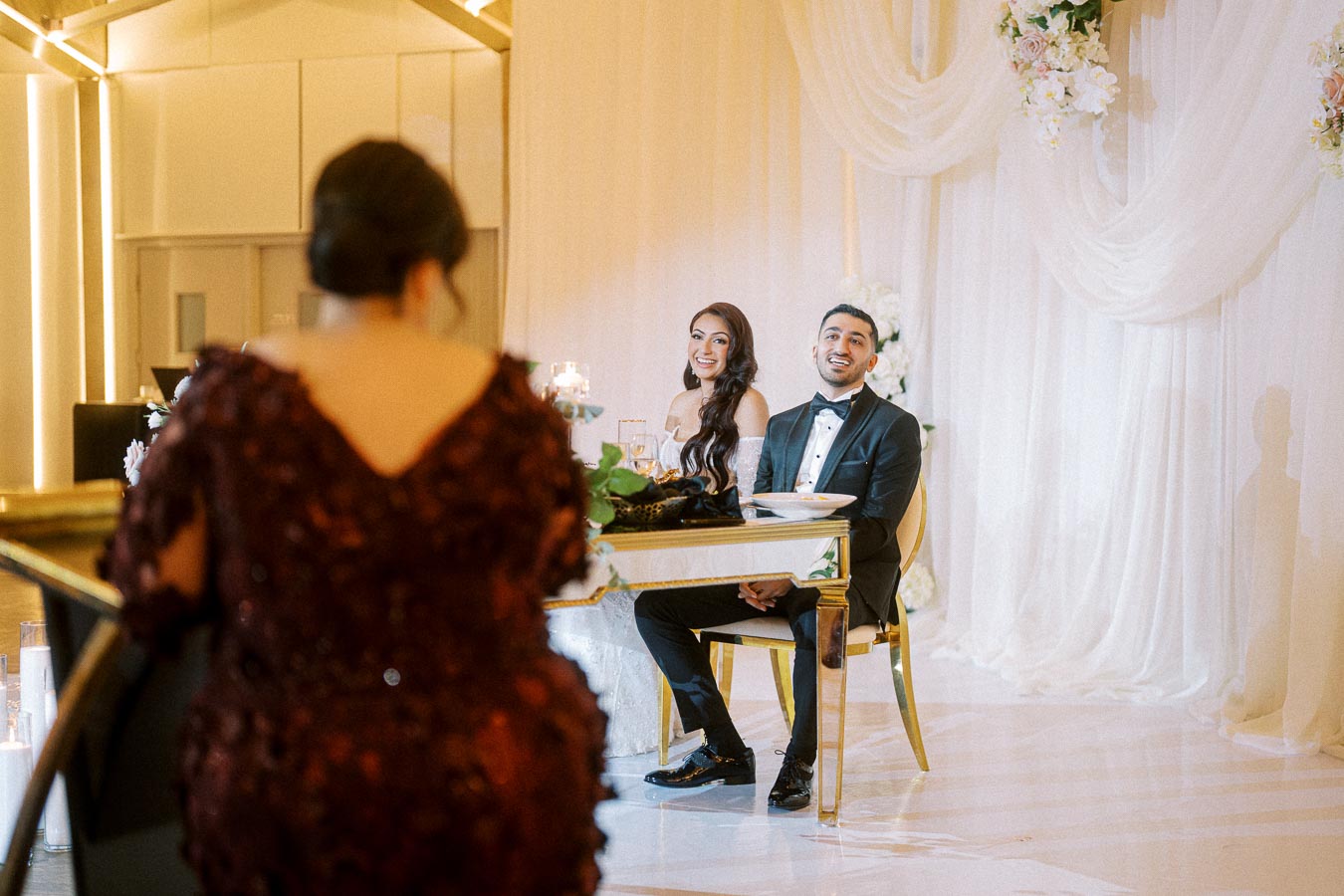 Elegant wedding reception with a bride and groom smiling at the head table, surrounded by white floral decorations and soft