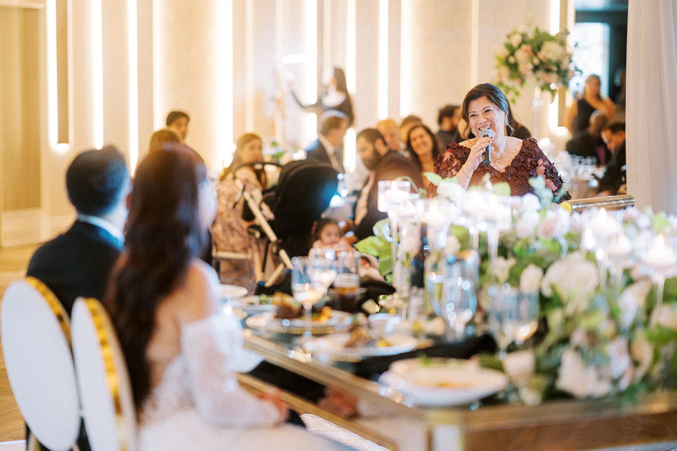Wedding reception scene with a woman in a maroon dress giving a speech at a beautifully decorated table. Guests are seated