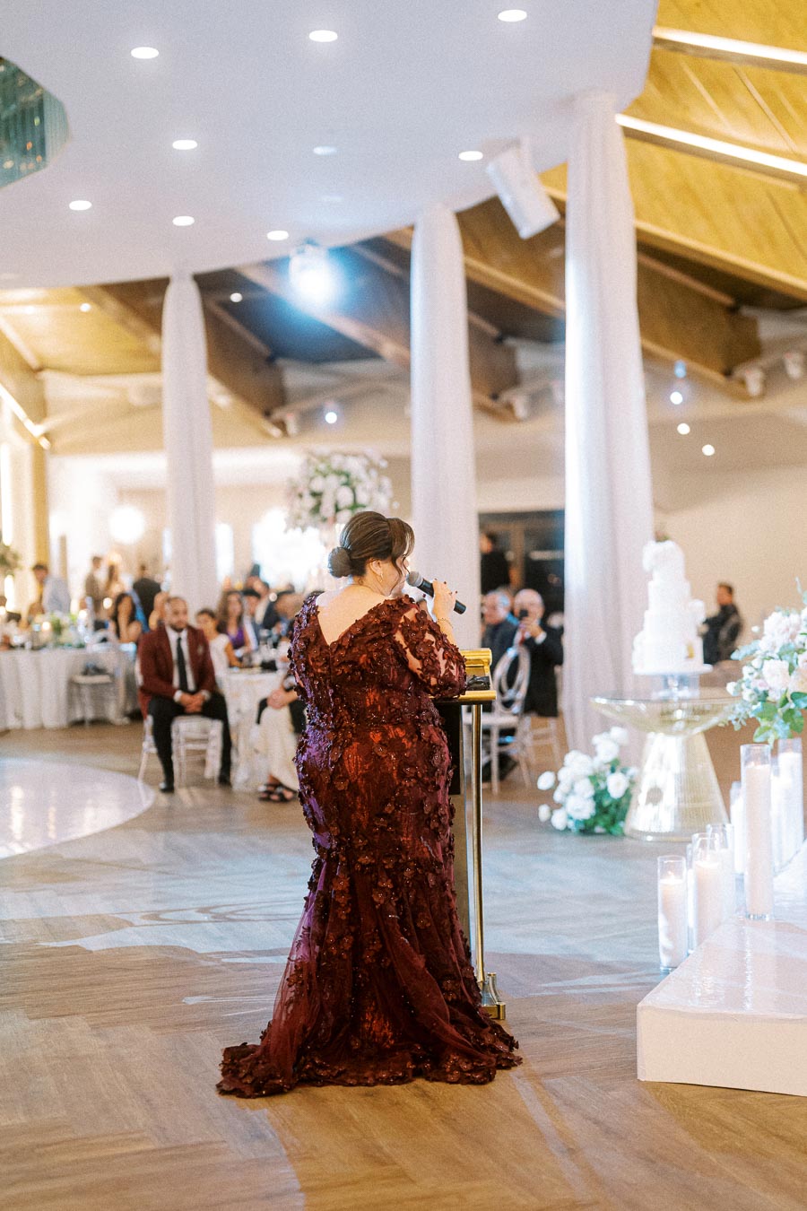 A woman in a burgundy dress gives a speech at a wedding reception, standing near a large white cake and elegant floral