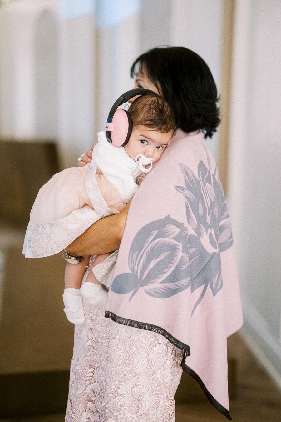 A woman holding a baby with pink noise-canceling earmuffs, wrapped in a floral-patterned pink blanket, in an indoor setting.
